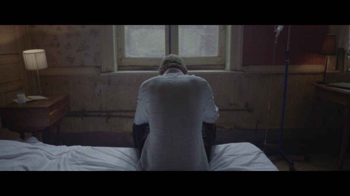 extrait du film post-apocalyptique Safeland. Un homme est assis de dos sur un lit. Il fait face à une fenêtre mais regarde le sol.